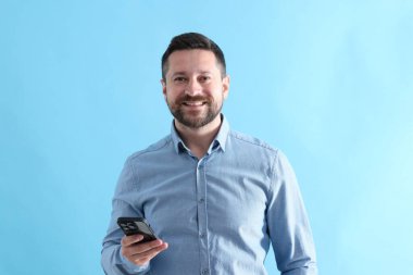 Akıllı telefonlu yakışıklı adam açık mavi arka planda gülüyor.