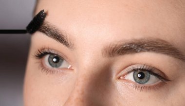 Genç bir kadın açık gri arka planda kaşlarını hortlakla fırçalıyor. Yakın plan.