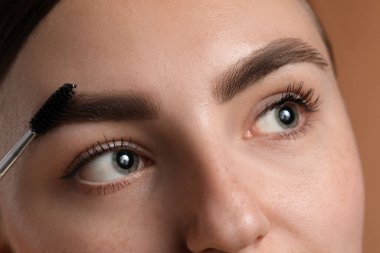 Kahverengi arka planda kaşlarını korkunç bir şekilde fırçalayan genç bir kadın.