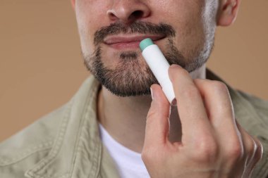 Açık kahverengi arka planda dudaklarına dudak kremi süren adam, yakın plan.