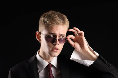 Siyah arka planda şık bir takım elbise ve güneş gözlüğü takan yakışıklı bir genç.