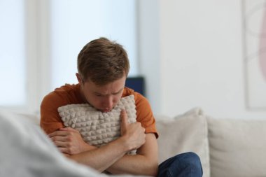 Üzgün adam evdeki kanepede yastığa sarılıyor.