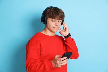 Açık mavi arka planda akıllı telefonu olan kulaklıklı tatlı bir genç çocuk.
