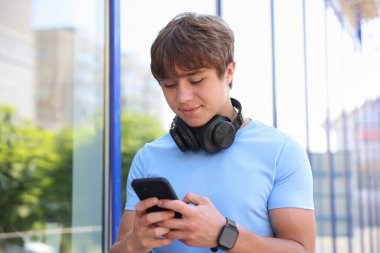 Kulaklığı ve akıllı telefonu olan sevimli genç çocuk binanın yanında.