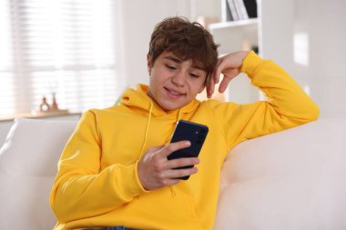 Evdeki kanepede akıllı telefon kullanan gülümseyen genç çocuk.