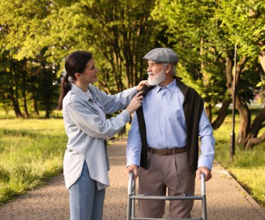 Gülümseyen bakıcı dışarıda yürüyen çerçeveyi kullanan yaşlı bir adamı destekliyor.