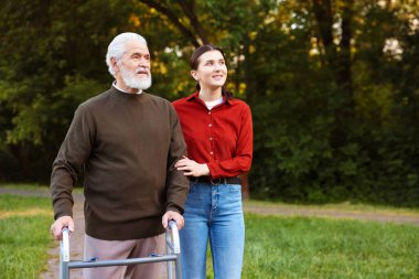 Gülümseyen bakıcı dışarıda yürüyen çerçeveyi kullanan yaşlı bir adamı destekliyor.