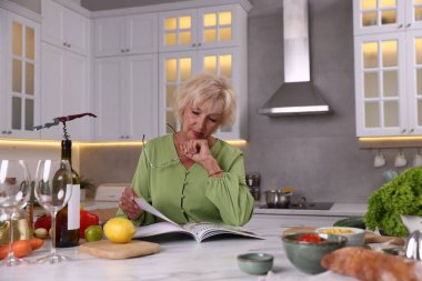 Mutfaktaki mermer masada gözlüklü yaşlı kadın dergisi okuyor.