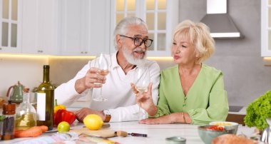 Yaşlı çift mutfakta yemek pişirirken beyaz şarap içiyor.