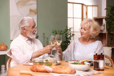 Yaşlı bir çift, akşam yemeği sırasında evde beyaz şarap bardaklarını tokuşturuyor.