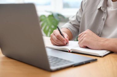 Metin yazarı dizüstü bilgisayar kullanıyor ve deftere yazıyor. Kapalı masa, yakın plan.