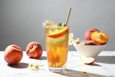 Bardakta buzlu şeftali çayı, taze meyve, nane ve beyaz masada papatya çiçeği.