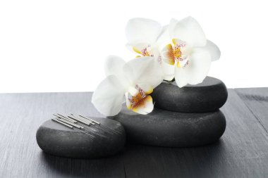Akupunktur iğneleri, orkide çiçekleri ve gri ahşap masadaki spa taşları beyaz arka planda, yakın plan