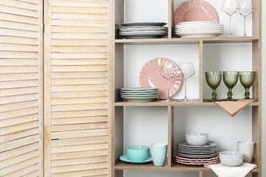 Beyaz duvarın yanında farklı bulaşık takımları ve gözlüklerle raf ünitesi.