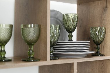 Kapalı alandaki yeşil duvarın yakınındaki ahşap raflarda farklı tabaklar ve bardaklar var.