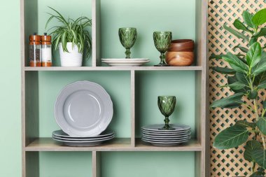Farklı bulaşık takımları, gözlükler ve yeşil duvarın yanında çiçek dekoru olan raf ünitesi.