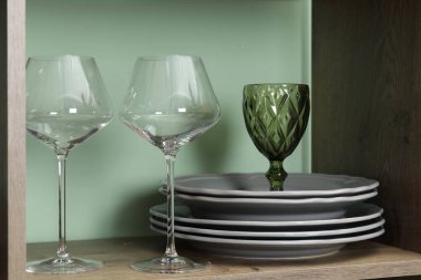 Kapalı alandaki yeşil duvarın yanındaki ahşap rafta farklı tabaklar ve bardaklar var.