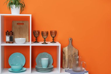 Farklı bulaşık takımları, fincanlar ve turuncu duvarın yanında çiçek dekoru olan raf ünitesi.