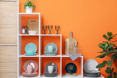 Farklı bulaşık takımları, fincanlar, bardaklar ve turuncu duvarın yanında çiçek dekoru olan raf ünitesi.