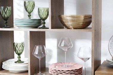 Beyaz duvarın yanında farklı bulaşık takımları ve gözlüklerle raf ünitesi.