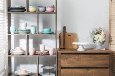 Beyaz duvarın yanında farklı bulaşık takımları, fincanlar ve çiçek dekoru olan raf ünitesi.