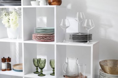 Beyaz duvarın yanında farklı bulaşık takımları, gözlük ve çiçek dekoru olan raf ünitesi.