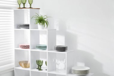 Beyaz duvarın yanında farklı bulaşık takımları, gözlük ve çiçek dekoru olan raf ünitesi.