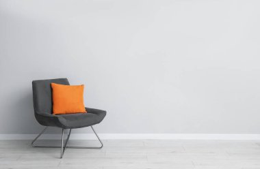 Açık gri duvarın yanında rahat bir koltuk ve turuncu yastık. Metin için boşluk