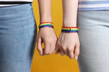 LGBT konsepti. Gökkuşağı bileklikli kadınlar sarı arka planda el ele tutuşuyorlar, yakın plan.