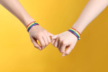 LGBT konsepti. Gökkuşağı bileklikli kadınlar sarı arka planda el ele tutuşuyorlar, yakın plan.