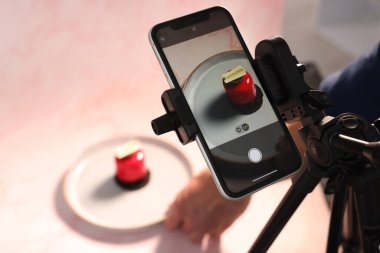 Adam stüdyodaki masada fotoşoplar için lezzetli tatlılarla kompozisyon hazırlıyor, akıllı telefon ve tripoda odaklanıyor. Profesyonel yemek fotoğrafçılığı.