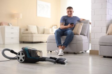 Evdeki koltukta akıllı telefonu olan adam, elektrik süpürgesine odaklan.