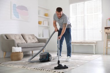 Evde süpürgeyle halı temizleyen bir adam var.