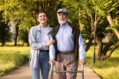 Gülümseyen bakıcı dışarıda yürüyen çerçeveyi kullanan yaşlı bir adamı destekliyor.