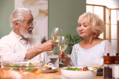 Yaşlı bir çift, akşam yemeği sırasında evde beyaz şarap bardaklarını tokuşturuyor.