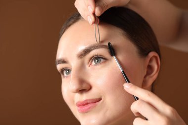Kozmetik uzmanı, kahverengi arka planda genç bir kadının kaşlarını yoluyor ve fırçalıyor.