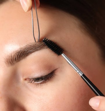 Kozmetik uzmanı genç bir kadının kaşlarını yoluyor ve fırçalıyor.