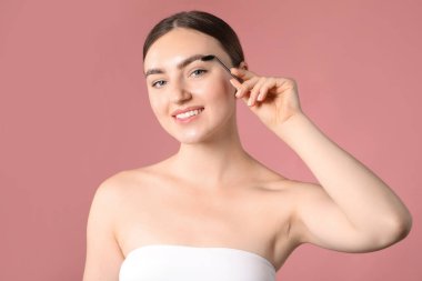 Genç bir kadın pembe arka planda kaşlarını hortlakla fırçalıyor.