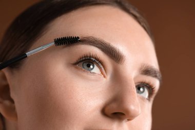 Kahverengi arka planda kaşlarını korkunç bir şekilde fırçalayan genç bir kadın.