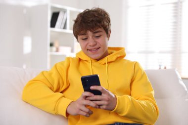Evdeki kanepede akıllı telefon kullanan gülümseyen genç çocuk.