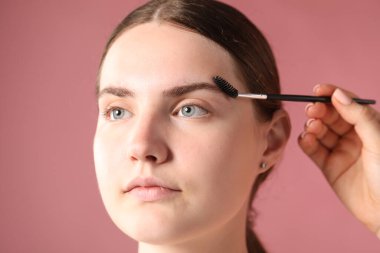 Kozmetik uzmanı genç kadının kaşlarını pembe arka planda hortlakla fırçalıyor.