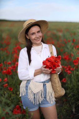 Şapkalı gülümseyen bir kadının portresi. Yaban çiçeği çayırında bir torba gelincik çiçeğiyle.
