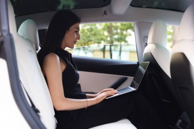 Modern arabada dizüstü bilgisayar kullanan bir kadın. Dışarıdan görünüyor.
