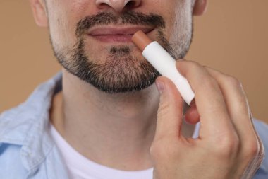 Açık kahverengi arka planda dudaklarına dudak kremi süren adam, yakın plan.