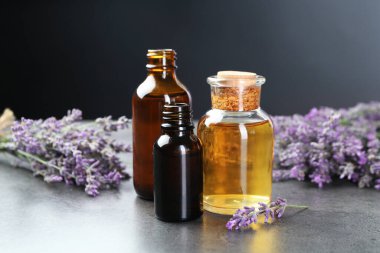 Şişelerdeki aromatik yağ ve gri desenli masadaki lavanta çiçekleri koyu arka planda, yakın plan.