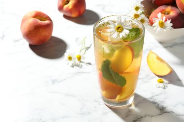 Lezzetli buzlu şeftali çayı bardakta, taze meyve, nane ve papatya çiçekleri beyaz masada, mesaj için yer var.