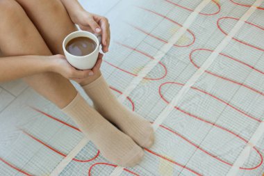 Bir fincan kahveli kadın sıcak zeminde oturuyor, yakın plan. Yeraltı ısıtma sistemi, çifte maruz kalma.