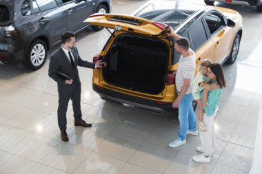 Elinde dosya olan satıcı salonda mutlu bir aileye yeni bir araba gösteriyor.