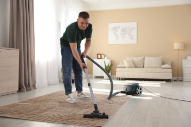 Evde süpürgeyle halı temizleyen bir adam var.