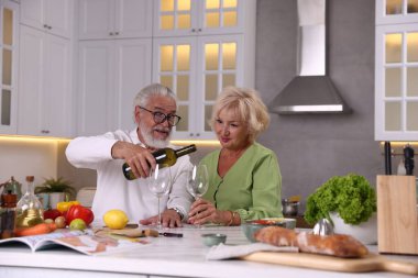 Yaşlı çift mutfakta yemek pişirirken beyaz şarap içiyor.